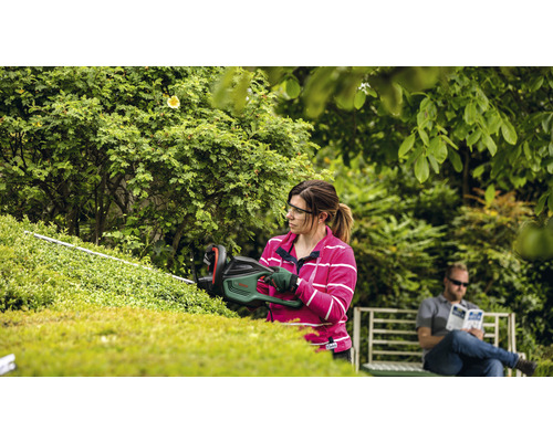 Frau schneidet Hecke mit Heckenschere im Garten, während ein Mann auf einer Bank sitzt