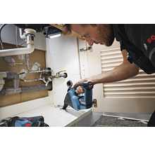Handwerker arbeitet mit einer Akku-Säbelsäge unter einem Waschbecken. Im Hintergrund ist eine Arbeitsleuchte zu sehen.
