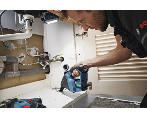 Handwerker arbeitet mit einer Akku-Säbelsäge unter einem Waschbecken. Im Hintergrund ist eine Arbeitsleuchte zu sehen.