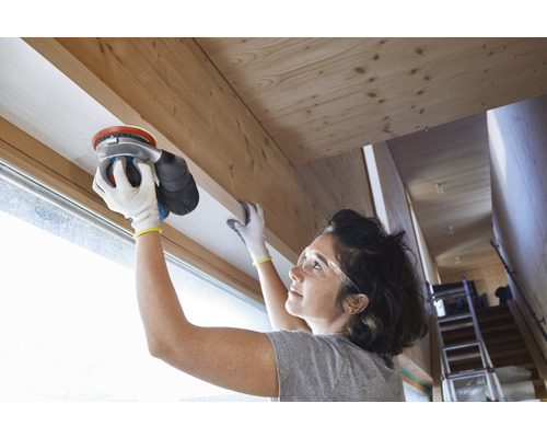 Eine Frau schleift einen hölzernen Fensterrahmen mit einem Exzenterschleifer.