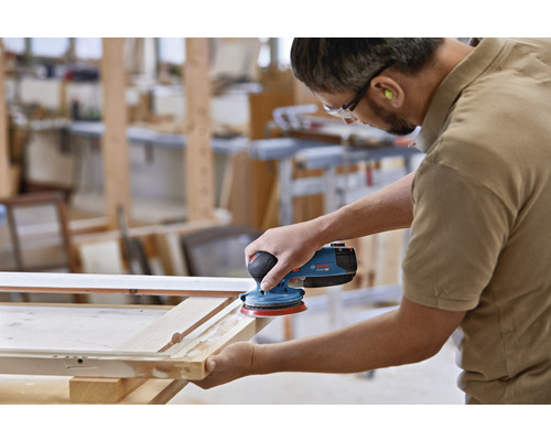 Ein Mann schleift mit einem Exzenterschleifer ein Holzstück in einer Werkstatt.