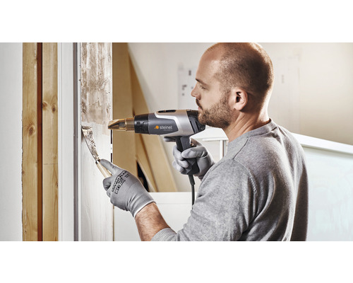 Ein Mann entfernt alte Farbe mit einem Heißluftfön und Spachtel von einem Fensterrahmen