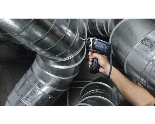 Eine Hand hält einen Akkuschlagschrauber vor einem Hintergrund aus Metallrohren.
