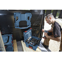 Ein Handwerker im Laderaum eines Transporters mit Werkzeugkoffer, montiertes Regalsystem und Akkugerät in der Hand.