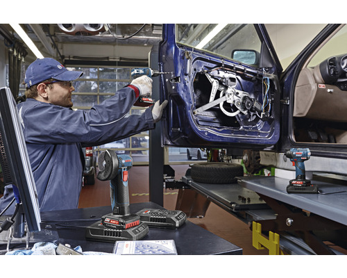 Ein Mann repariert eine Autotür mit einem Akku-Bohrschrauber. Auf dem Tisch liegen ein Winkelschleifer und ein Akkupack.
