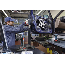 Werkstatt-Szene mit einem Handwerker, der mit einem Akku-Bohrschrauber an einer Autotür arbeitet, im Vordergrund Akku-Winkelschleifer und Akku-Schlagschrauber.