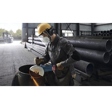 Ein Mann mit Schutzkleidung bearbeitet ein Metallrohr mit einem Winkelschleifer in einer Werkstatt.