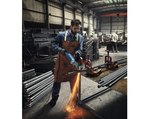 Ein Mann mit Schutzbrille und Lederschürze schneidet mit einem Winkelschleifer ein Metallrohr in einer Werkstatt.