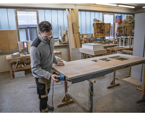 Ein Handwerker bearbeitet eine Holztür mit einem Elektrowerkzeug in einer Werkstatt.