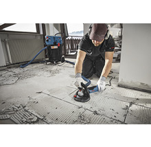 Ein Mann verwendet einen Betonschleifer auf einem Boden, der mit einem Staubsauger verbunden ist. Bosch Logo.