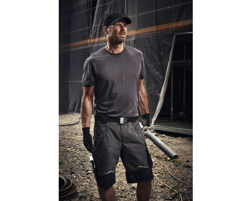 Handwerker trägt Arbeitskleidung mit Shorts, T-Shirt, Gürtel und Handschuhen auf einer Baustelle