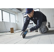 Handwerker schneidet Bodenbelag mit einem Elektrowerkzeug in einem Raum. Im Hintergrund sind Fliesen zu sehen.
