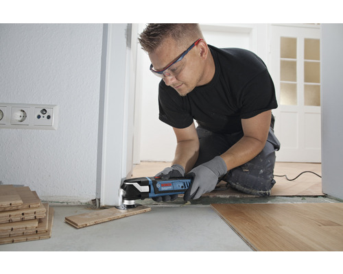 Ein Handwerker verwendet ein Multifunktionswerkzeug zum Zuschneiden eines Holzstücks, um es an eine Tür anzupassen. Das Werkzeug ist blau und schwarz.