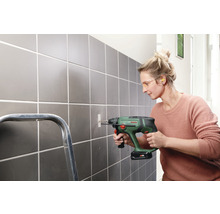 Eine Frau bohrt mit einer Bohrmaschine ein Loch in eine geflieste Wand. Sie trägt eine Schutzbrille und Ohrstöpsel.