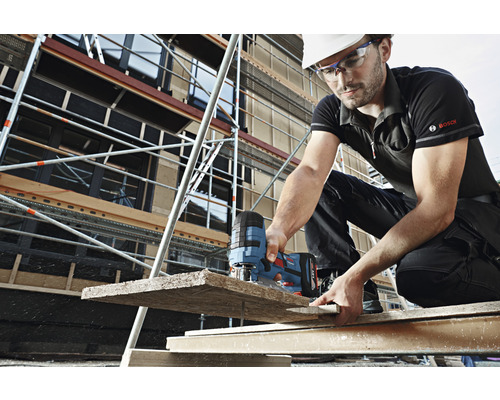 Ein Bauarbeiter mit Schutzhelm und Brille sägt ein Holzbrett mit einer Stichsäge auf einer Baustelle. Im Hintergrund befindet sich ein Baugerüst.