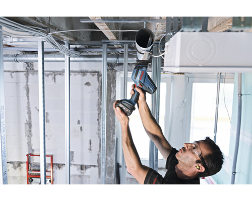 Ein Handwerker montiert mit einem Akku-Bohrschrauber ein Rohr an einer Decke in einem Rohbau.