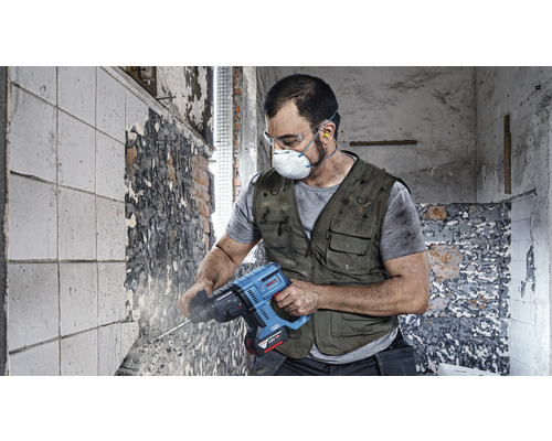Ein Arbeiter mit Schutzbrille, Maske und Gehörschutz verwendet einen Bohrhammer an einer Wand.