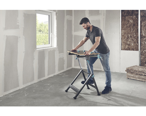 Mann arbeitet mit einer Werkbank in einem Raum mit Trockenbauwänden und einem Fenster.