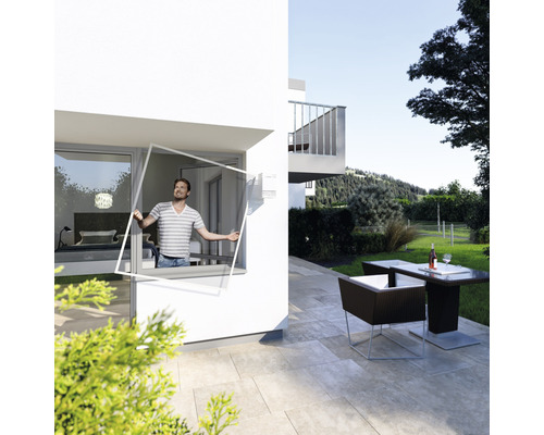 Ein Mann montiert einen Insektenschutzrahmen an einem Fenster eines modernen Hauses, während im Hintergrund ein Gartensitzplatz mit Tisch und Stühlen zu sehen ist.
