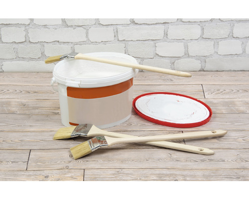 Eimer mit Farbe, Farbpinsel und Farbabstreifer auf einem Holzboden vor einer weißen Ziegelwand