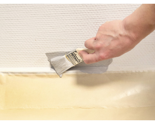 Eine Person streicht mit einem Pinsel eine Wand entlang einer Abdeckbandlinie.