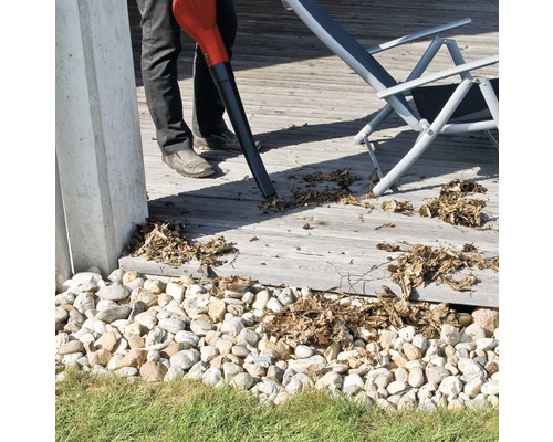 Eine Person verwendet einen Laubbläser, um Blätter von einer Holzterrasse zu entfernen. Im Hintergrund ist ein Liegestuhl zu sehen.