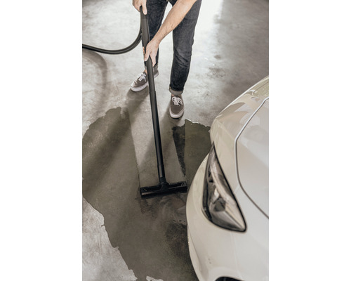 Eine Person reinigt mit einem Nass Trockensauger eine Wasserlache neben einem Auto auf einem Garagenboden.