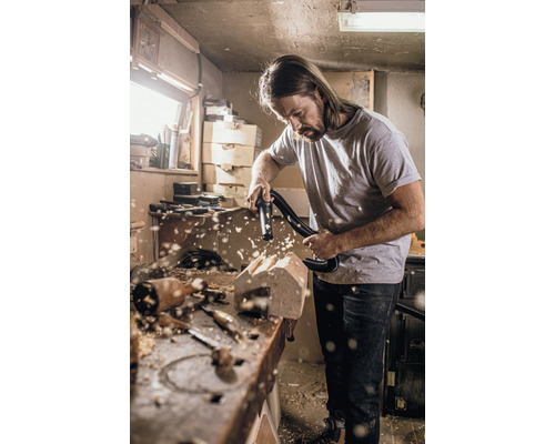 Mann saugt Holzstaub von einer Holzskulptur mit einem Staubsauger in einer Werkstatt.
