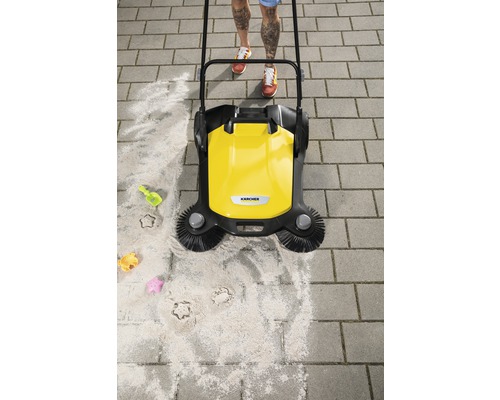 Gelbe Kehrmaschine im Einsatz auf Pflastersteinen mit Sand und Kinderspielzeug.