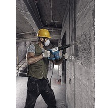Ein Bauarbeiter mit Schutzhelm und Staubmaske verwendet einen Bohrhammer an einer Betonwand.