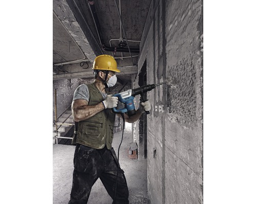 Ein Bauarbeiter mit Schutzhelm und Staubmaske verwendet einen Bohrhammer an einer Betonwand.