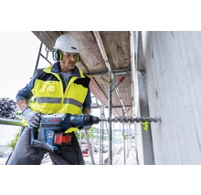 Bauarbeiter bohrt mit Bohrhammer ein Loch in eine Betonwand auf einem Baugerüst. Er trägt Schutzkleidung und einen Helm.