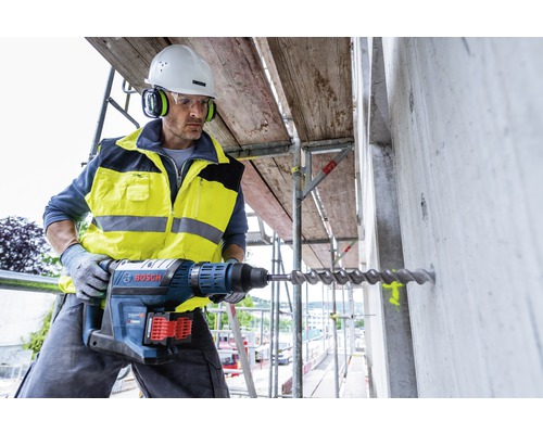 Bauarbeiter bohrt mit Bohrhammer ein Loch in eine Betonwand auf einem Baugerüst. Er trägt Schutzkleidung und einen Helm.