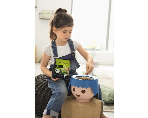 Mädchen befüllt Pflanzgefäß in Form eines Gesichts mit Rasensamen, hält Samenumschlag, Bio Siegel