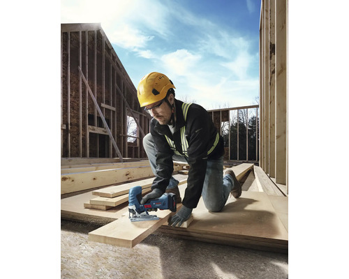 Ein Bauarbeiter mit Schutzhelm und Schutzbrille sägt ein Holzbrett mit einer Stichsäge auf einer Baustelle.