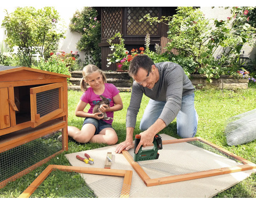 Vater und Tochter bauen einen Hasenstall im Garten mit einem Tacker, Holz und Drahtgitter.