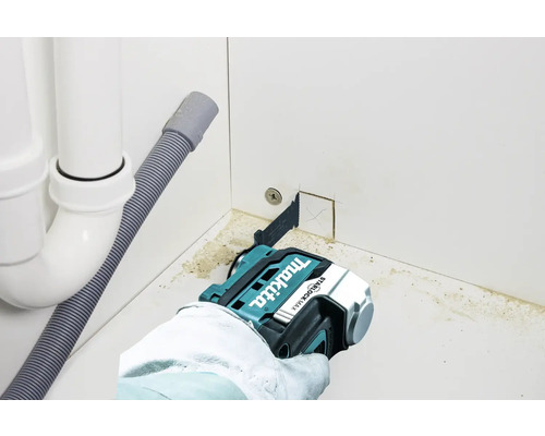 Eine Hand mit Handschuh verwendet eine oszillierende Multifunktionssäge der Marke Makita, um ein Loch in eine weiße Küchenwand zu sägen.