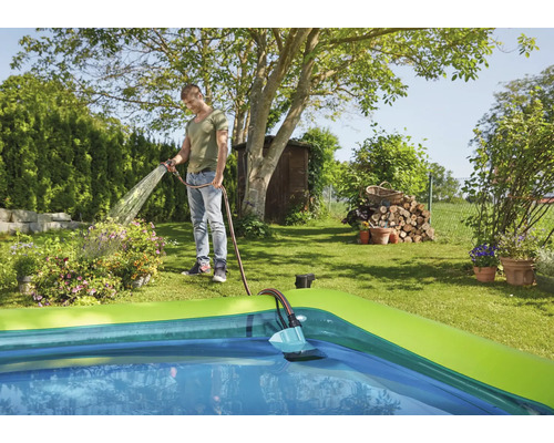 Mann bewässert Pflanzen mit Gartenschlauch neben einem Pool im Garten.