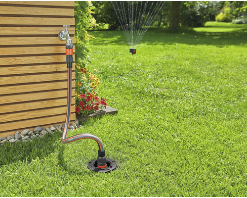 Gartenszene mit Wasserhahn, Gartenschlauch und Rasensprenger auf Rasenfläche.