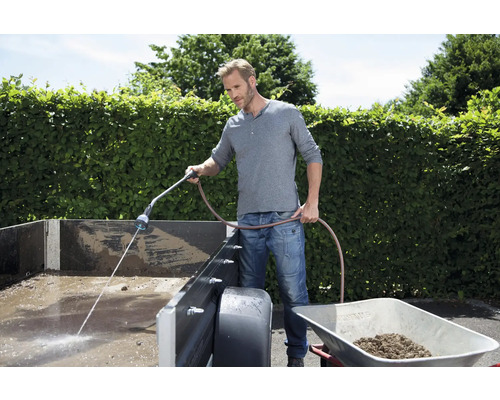 Ein Mann reinigt einen Anhänger mit einem Gartenschlauch und einer Gartenspritze. Eine Schubkarre steht daneben.