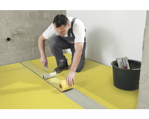 Handwerker bringt mit einer Farbwalze eine Abdichtungsbahn auf den Boden auf. Im Hintergrund ist ein Eimer mit Werkzeugen zu sehen.