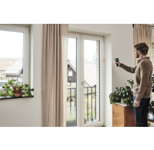 Mann misst mit einem Infrarot Thermometer die Temperatur an einem Fenster in einem hellen Wohnraum.