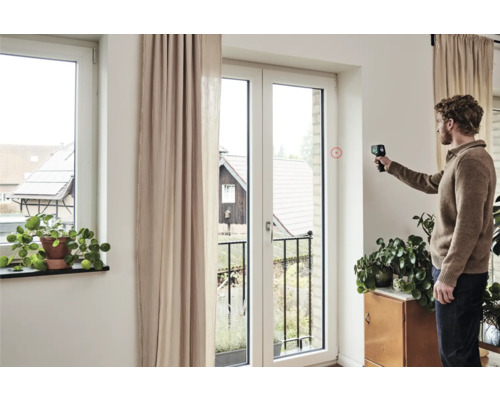 Mann misst mit einem Infrarot Thermometer die Temperatur an einem Fenster in einem hellen Wohnraum.