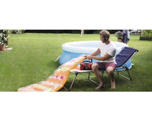 Mann pumpt Luftmatratze mit einer Luftpumpe im Garten auf, im Hintergrund ein Pool