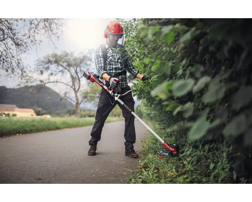 Eine Person trimmt eine Hecke mit einer Akku-Grasschere, die mit einem Tragegurt getragen wird. Die Person trägt Schutzkleidung.