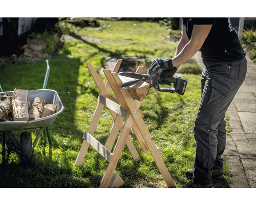 Eine Person sägt mit einer Akku-Kettensäge ein Stück Holz auf einem Holzsägebock. Eine Schubkarre mit Holz befindet sich im Hintergrund.