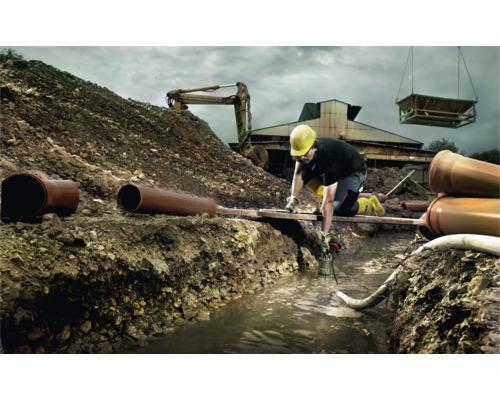 Bauarbeiter bei der Arbeit in einem Graben mit Rohren, Bagger und Kran im Hintergrund.