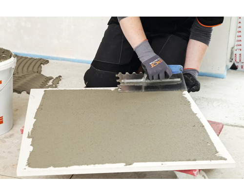 Eine Person trägt mit einer Zahnspachtel Kleber auf eine Fliese auf.