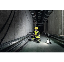 Ein Arbeiter mit Schutzhelm und Schutzweste bohrt mit einer Bohrmaschine in eine Betonwand, beleuchtet von einem Arbeitsstrahler.