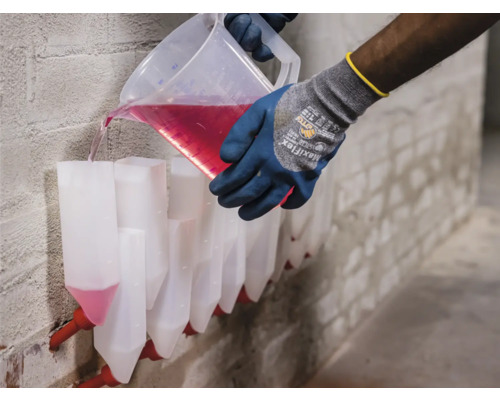 Eine Person gießt mit Handschuhen eine rote Flüssigkeit aus einem Messbecher in eine Reihe von Injektionskanülen an einer Wand.